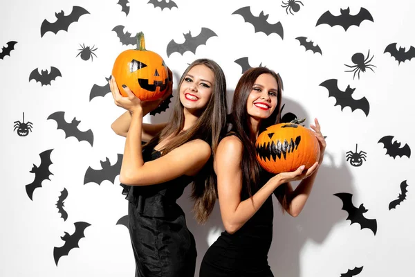 Zwei charmante brünette Frauen in schwarzen Kleidern mit rotem Lippenstift halten Halloween-Kürbisse auf weißem Hintergrund mit Fledermäusen und Spinnen — Stockfoto