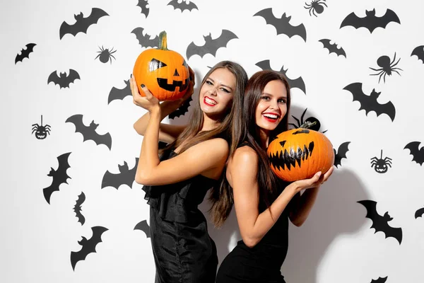Twee charmante brunette vrouw in zwarte jurken met rode lippenstift houden Halloween pompoenen op een witte achtergrond met vleermuizen en spinnen — Stockfoto