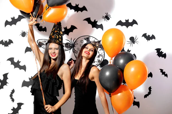 Dos mujeres morenas en vestidos negros y sombreros de brujas se divierten con globos negros y naranjas sobre un fondo blanco con murciélagos negros —  Fotos de Stock