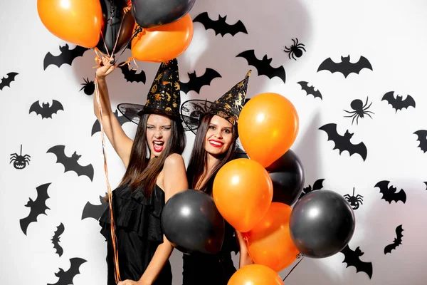 两个穿着黑色礼服和女巫帽子的黑发妇女在白色背景上与黑色蝙蝠的黑色和橙色气球一起享受乐趣 — 图库照片