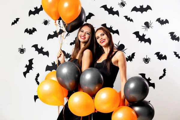 Twee jonge vrouwen in zwarte jurken en hoeden van de witchs veel plezier met zwart en oranje ballonnen op een witte achtergrond met zwarte vleermuizen — Stockfoto