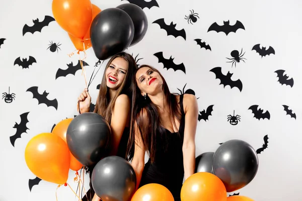 Zwei junge Frauen in schwarzen Kleidern und Hexenhüten haben Spaß mit schwarzen und orangefarbenen Luftballons auf weißem Hintergrund mit schwarzen Fledermäusen — Stockfoto