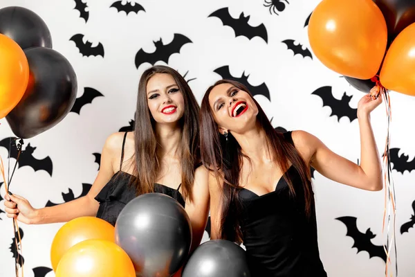 Donne brune in abiti neri e cappelli streghe si divertono con palloncini neri e arancioni su uno sfondo con pipistrelli neri. Halloween — Foto Stock