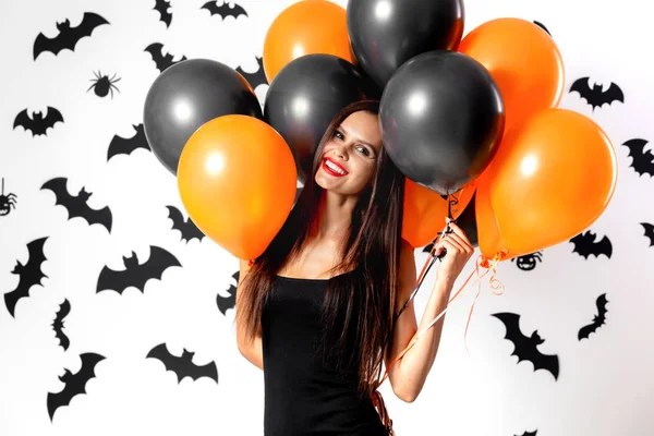 Ursnygg brunett kvinna i svart klänning innehar svart och orange ballonger på en vit bakgrund med svarta fladdermöss och spindlar. Halloween — Stockfoto
