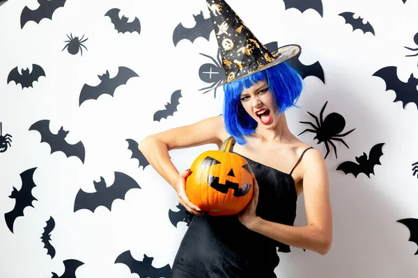 Lustige junge Frau mit blauer Perücke und Hexenhut hält Halloween-Kürbis auf weißem Hintergrund mit schwarzen Fledermäusen und Spinnen — Stockfoto