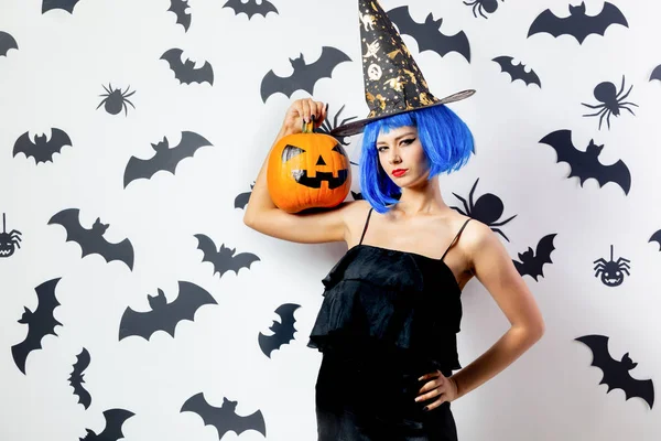 Cadılar Bayramı Balkabağı siyah yarasalar ve örümcekler beyaz bir arka plan üzerinde mavi bir peruk ve witchs şapkalı komik genç kadın tutar