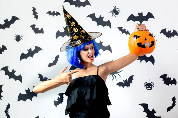 Vicces fiatal nő, egy kék paróka és witchs kalap tartja Halloween tök fehér háttér, fekete denevérek és a pókok — Stock Fotó