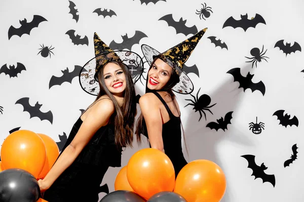 Dos encantadoras mujeres jóvenes con sombreros de brujas sostienen globos negros y naranjas sobre un fondo blanco con murciélagos negros. Confetti alrededor —  Fotos de Stock