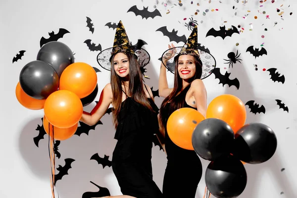 Dos hermosas mujeres con sombreros de brujas sonríen y sostienen globos negros y naranjas sobre un fondo de Halloween. Confetti alrededor —  Fotos de Stock