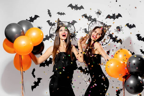 Zwei hinreißende Frauen mit Hexenhüten lächeln und halten schwarze und orangefarbene Luftballons auf einem Halloween-Hintergrund. Konfetti um sich — Stockfoto