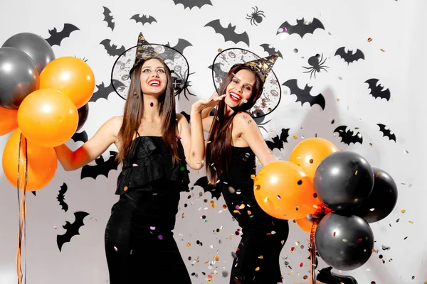 Dvě nádherné ženy v čarodějnice klobouky úsměv a podržte černé a oranžové balónky na pozadí Halloween. Konfety a okolí — Stock fotografie