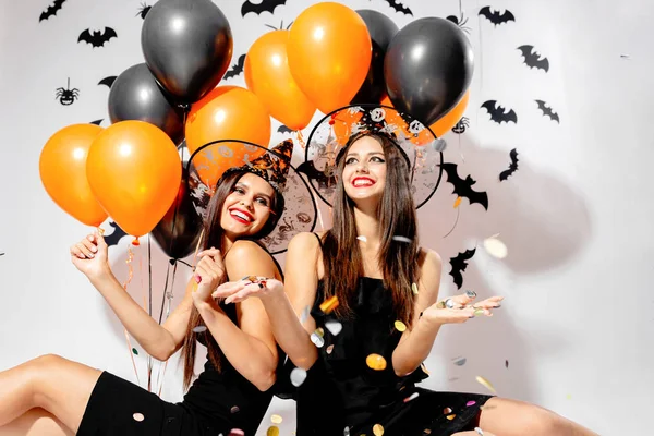 Dos mujeres morenas con sombreros de brujas sonríen y sostienen globos negros y naranjas. Confetti alrededor. Halloween — Foto de Stock