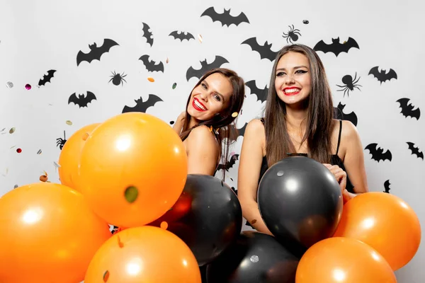Due donne brune in abiti neri sorridono e tengono palloncini neri e arancioni. Confetti in giro. Halloween — Foto Stock
