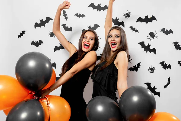 Ursnygg brunett kvinna i svart klänning innehar svart och orange ballonger på en vit bakgrund med svarta fladdermöss och spindlar. Halloween — Stockfoto