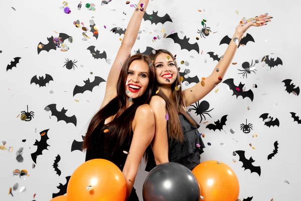 Dvě půvabné ženy v čarodějnice klobouky bavit s konfety a podržte černé a oranžové balónky na bílém pozadí s černým netopýři a pavouci. — Stock fotografie