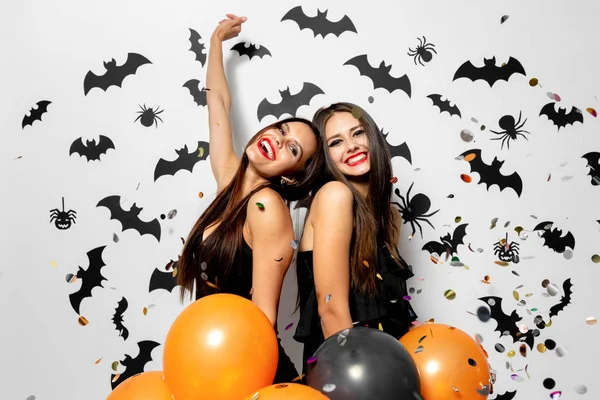 Zwei charmante Frauen mit Hexenhüten amüsieren sich mit Konfetti und halten schwarze und orangefarbene Luftballons vor weißem Hintergrund mit schwarzen Fledermäusen und Spinnen. — Stockfoto