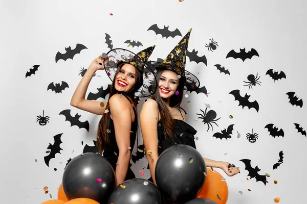 Två charmiga kvinnor i häxor hattar har kul med konfetti och håll svart och orange ballonger på en vit bakgrund med svarta fladdermöss och spindlar. — Stockfoto