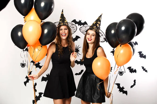 Due belle ragazze brune vestite con abiti e cappelli neri tengono palloncini neri e arancioni. Halloween — Foto Stock