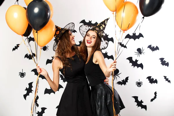 Zwei schöne brünette Mädchen in schwarzen Kleidern und Hexenhüten halten schwarze und orangefarbene Luftballons in der Hand. halloween — Stockfoto