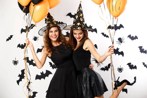 两个穿着黑色礼服和女巫帽的漂亮黑发女孩拿着黑色和橙色的气球。万圣节 — 图库照片