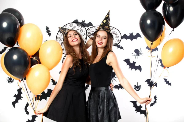 Zwei brünette Frauen in schwarzen Kleidern und Hexenhüten halten schwarze und orangefarbene Luftballons mit Fledermäusen an der Wand. Halloween-Party — Stockfoto