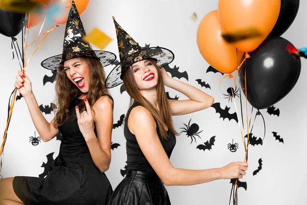 Due ragazze brune in abiti neri e cappelli da strega si divertono con palloncini sullo sfondo del muro con i pipistrelli. Halloween — Foto Stock