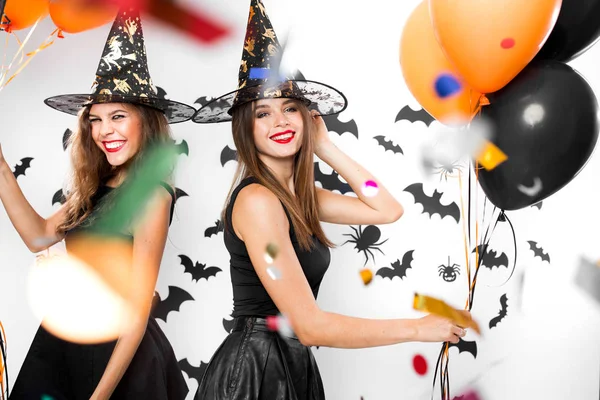 Zwei brünette Mädchen in schwarzen Kleidern und Hexenhüten amüsieren sich mit Luftballons und Konfetti. Halloween-Party — Stockfoto