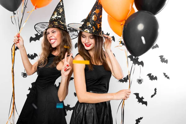 Twee brunette meisjes in zwarte jurken en hoeden van de heks hebben plezier met ballonnen en confetti. Halloweenfeest — Stockfoto