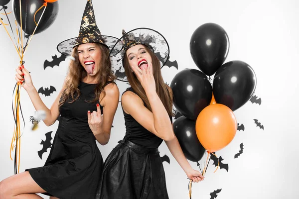 Zwei brünette Mädchen in schwarzen Kleidern und Hexenhüten haben Spaß mit Luftballons auf dem Hintergrund der Wand mit Fledermäusen. halloween — Stockfoto