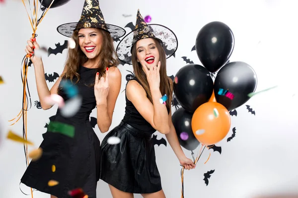 Halloween party. Two gorgeous girls in black dresses and witch hats have fun with balloons and confetti. — Stock Photo, Image