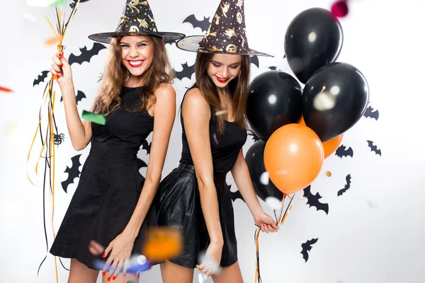 Halloween-feest. Twee beeldige meiden in zwarte jurken en hoeden van de heks hebben plezier met ballonnen en confetti. — Stockfoto