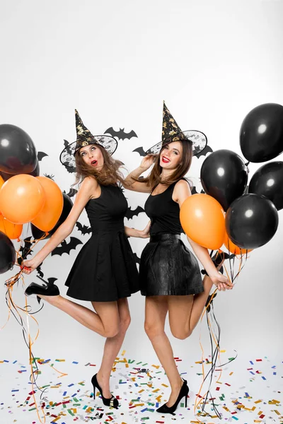 Zwei hinreißende Frauen in schwarzen Kleidern, Hexenhüten und High Heels halten schwarze und orangefarbene Luftballons in der Hand. Halloween-Party — Stockfoto
