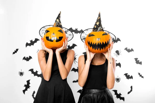 Zwei junge Frauen in schwarzen Kleidern und Hexenhüten halten sich Halloween-Kürbisse ins Gesicht — Stockfoto