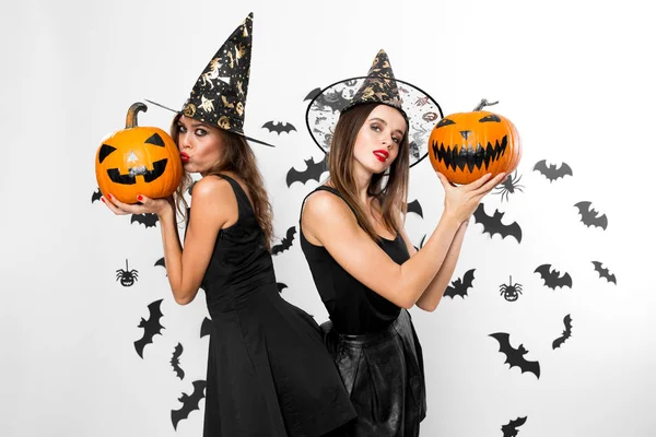 Zwei brünette Mädchen in schwarzen Kleidern und Hexenhüten halten Halloween-Kürbisse auf dem Hintergrund der Wand mit Fledermäusen. — Stockfoto