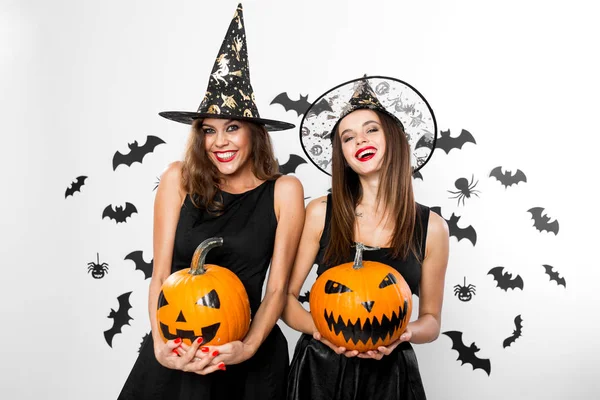 Zwei brünette Mädchen in schwarzen Kleidern und Hexenhüten halten Horror-Halloween-Kürbisse mit Fledermäusen im Hintergrund — Stockfoto