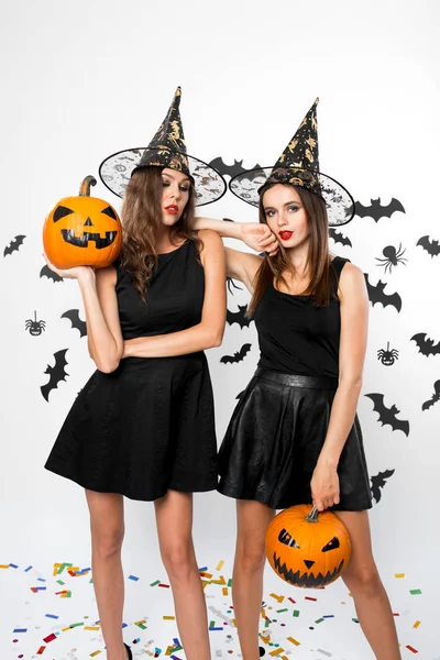 Due bellissime ragazze che indossano abiti neri e cappelli da strega tengono zucche di Halloween sullo sfondo con i pipistrelli — Foto Stock