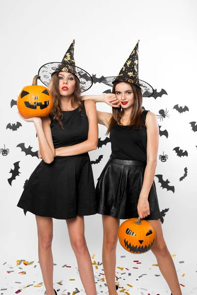 Due bellissime ragazze che indossano abiti neri e cappelli da strega tengono zucche di Halloween sullo sfondo con i pipistrelli — Foto Stock