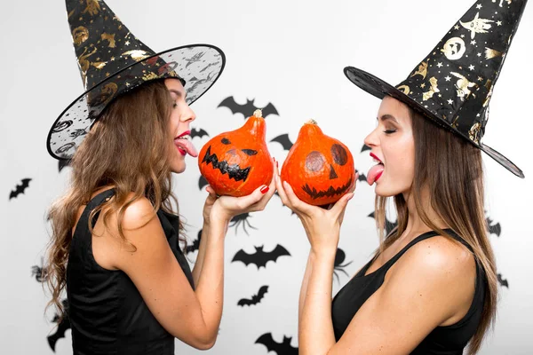 Due belle ragazze brune in cappelli da strega neri tengono le zucche di Halloween nelle loro mani e mostrano le loro lingue — Foto Stock