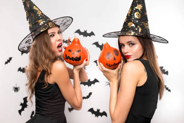 Dos hermosas chicas en vestidos negros y sombreros de brujas sostienen calabazas de halloween en sus manos en el fondo con murciélagos —  Fotos de Stock