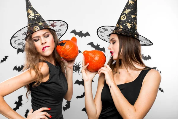 Två brunetter i svarta klänningar och häxa hattar grimas och håller Halloweenpumpor i sina händer — Stockfoto