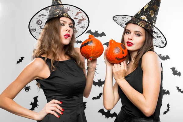 Dos chicas morenas en vestidos negros y sombreros de brujas hacen muecas y sostienen calabazas de halloween en sus manos —  Fotos de Stock