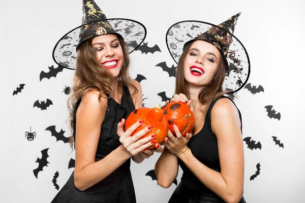 Due ragazze brune in abiti neri e cappelli da strega tengono le zucche di Halloween nelle loro mani e sorridono — Foto Stock