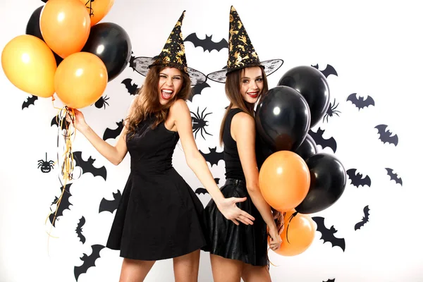 Due donne brune in abiti neri e cappelli da strega tengono palloncini neri e arancioni sullo sfondo del muro con i pipistrelli. Festa di Halloween — Foto Stock