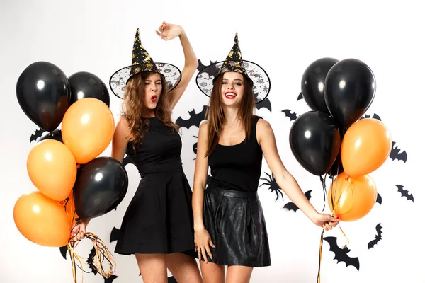 Dos mujeres morenas con vestidos negros y sombreros de brujas sostienen globos negros y naranjas en el fondo de la pared con murciélagos. Fiesta de Halloween —  Fotos de Stock