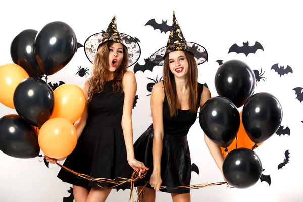 Två brunett kvinnor i svarta klänningar och häxa hattar innehar svart och orange ballonger på bakgrunden av väggen med fladdermöss. Halloween-fest — Stockfoto