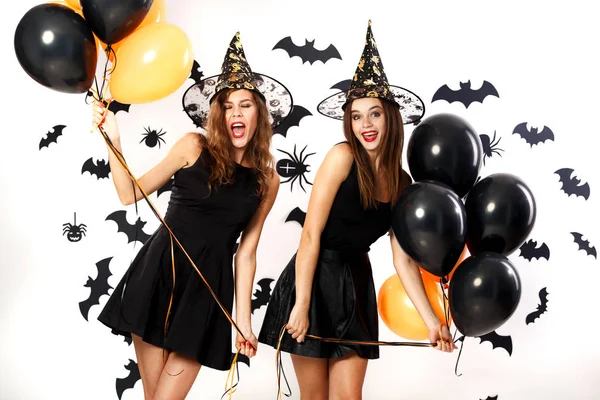 Twee mooie vrouwen in zwarte jurken en hoeden van de heks hebben plezier met zwart en oranje ballonnen. Halloweenfeest — Stockfoto