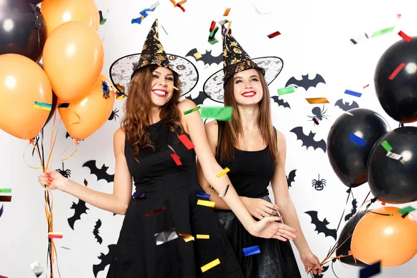 Due belle ragazze in abiti neri e cappelli da strega si divertono con palloncini neri e arancioni e confetti.Halloween party — Foto Stock