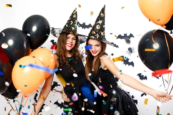 Zwei schöne Mädchen in schwarzen Kleidern und Hexenhüten amüsieren sich mit schwarzen und orangefarbenen Luftballons und Konfetti. Halloween-Party — Stockfoto
