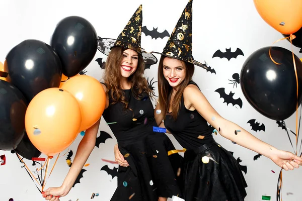 Zwei schöne Mädchen in schwarzen Kleidern und Hexenhüten amüsieren sich mit schwarzen und orangefarbenen Luftballons und Konfetti. Halloween-Party — Stockfoto