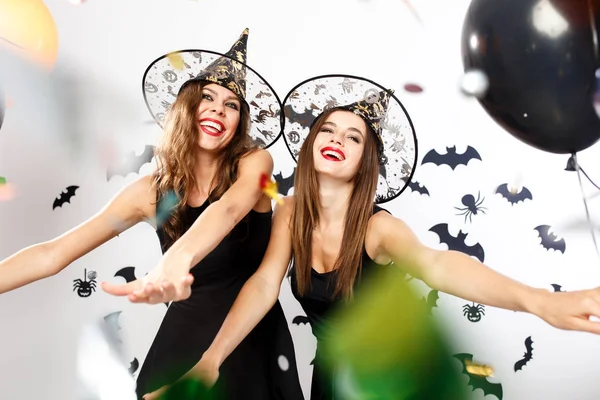 黒のドレスや魔女の帽子で 2 つの美しい女の子がバットで壁の背景に風船と紙吹雪楽しい時を過す — ストック写真